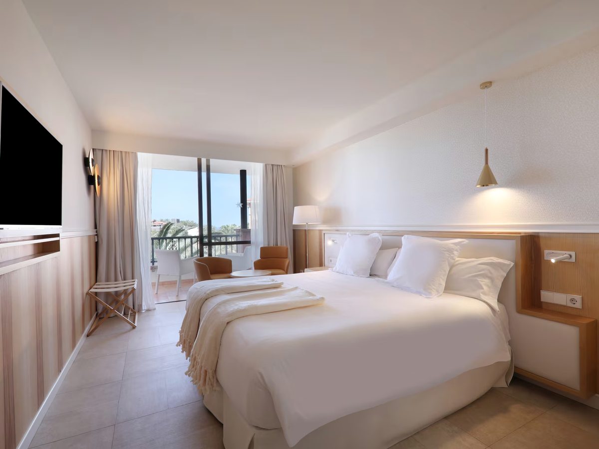 A guest room with twin beds pushed together, a wood headboard, a wall-mounted TV and sliding doors onto a balcony with a sea view.