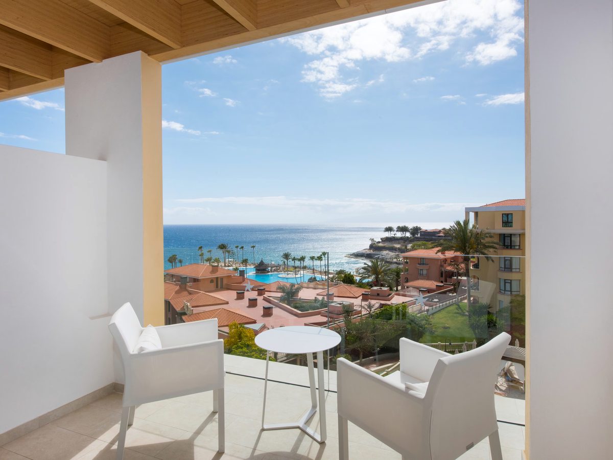 A covered balcony with a round table and white chairs above terracotta roofs, palms, the pools and the sea.