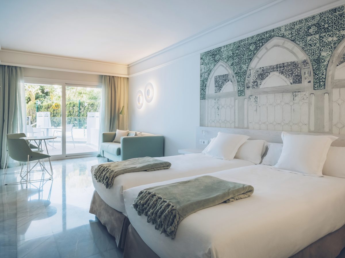 A twin room with a patterned tiled headboard, a small sofa and a table, and sliding glass doors onto a planted terrace.