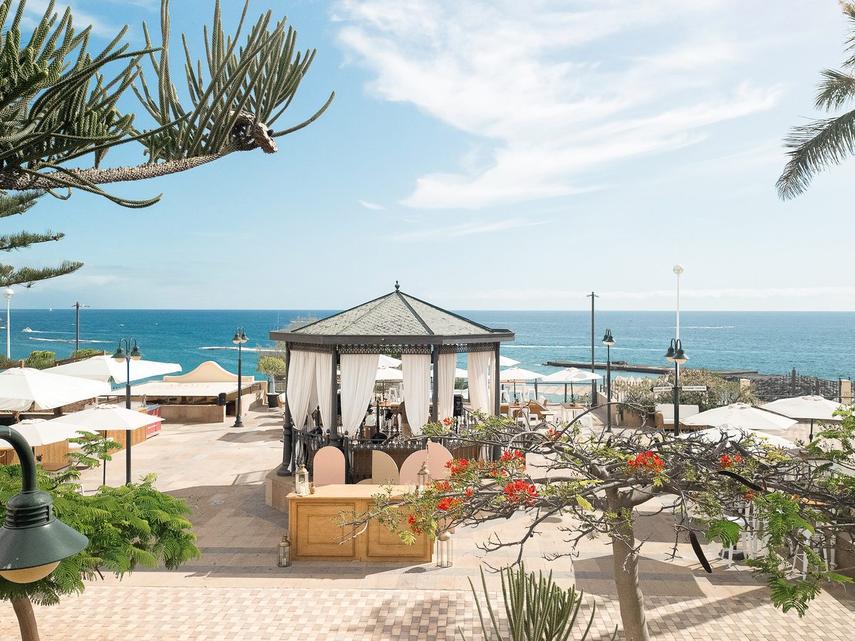 A garden terrace with a white gazebo, paved paths, white parasols and tropical planting beside the sea.