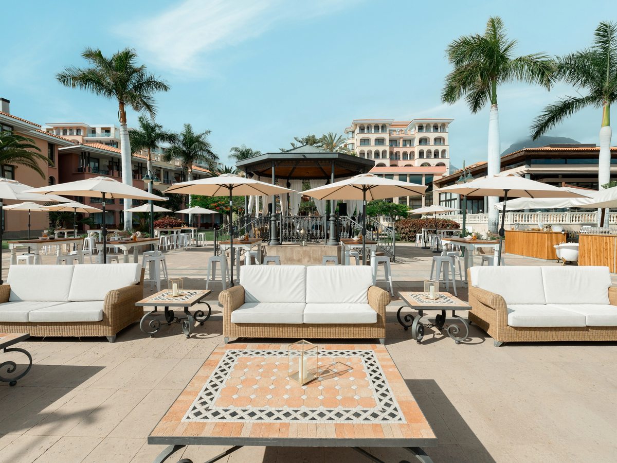 A terrace beside the pool with white sofa seating, low tiled tables and parasols among palms, the resort buildings behind.