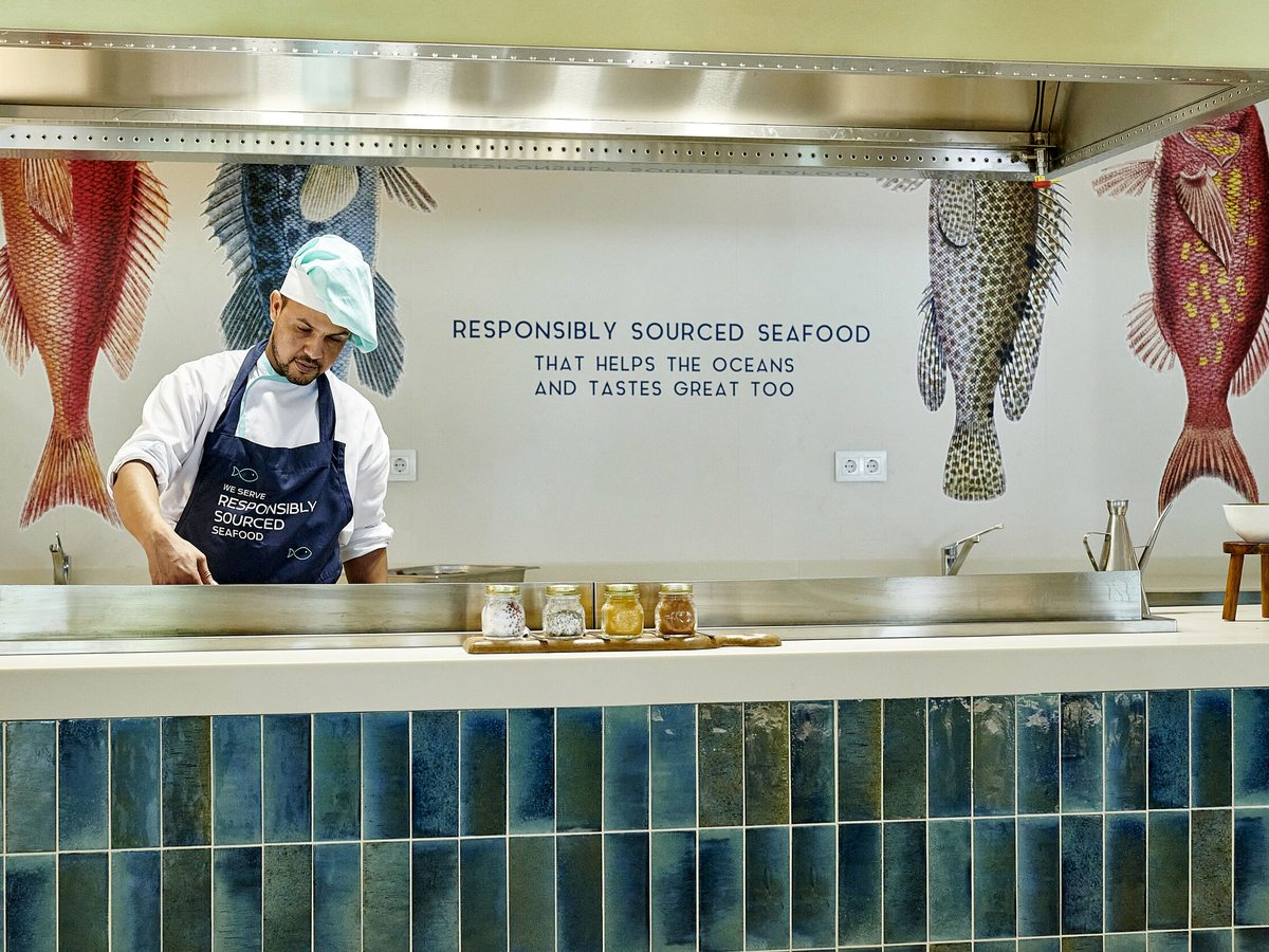 A chef in whites cooking at a blue-tiled buffet counter beneath a wall reading responsibly sourced seafood.