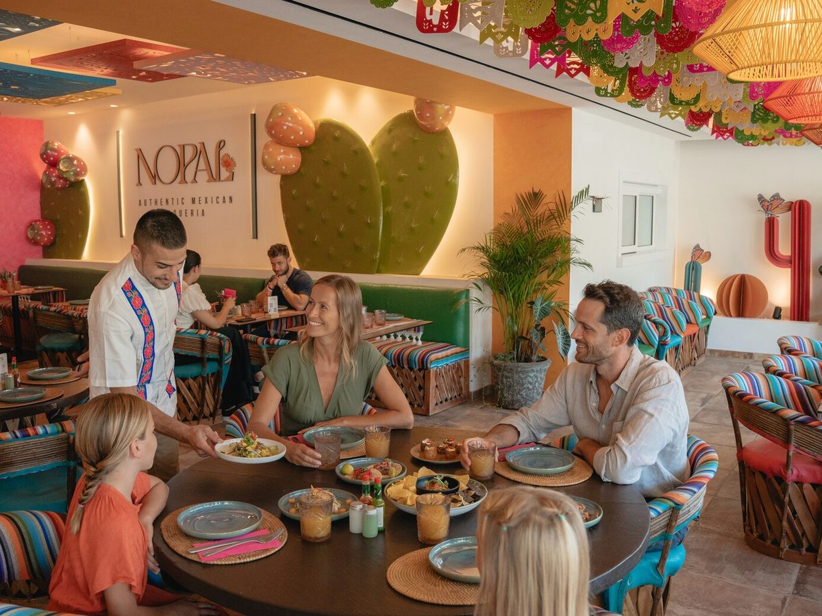 Families eating at round tables in the Nopal restaurant, a green cactus-motif wall and striped chairs behind them.