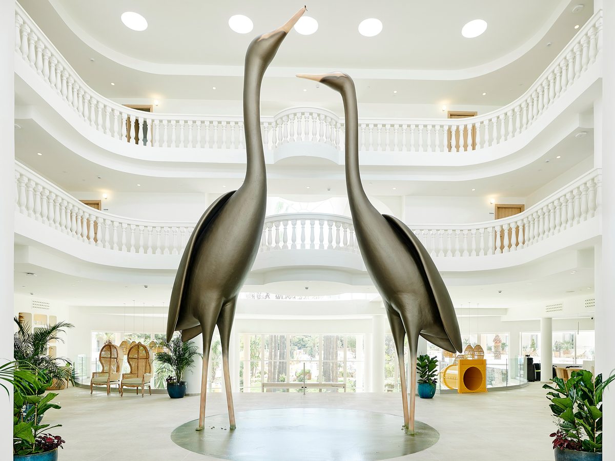 A bright white multi-storey lobby atrium with curved balconies and tall metal crane sculptures at its centre.