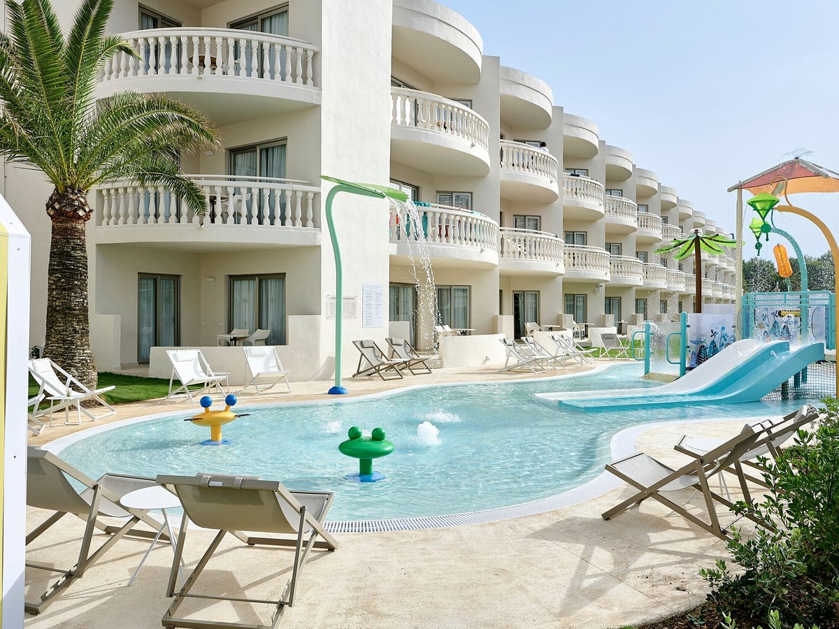 A shallow curved children's splash pool with a small blue water slide and frog-shaped water jets, sunloungers set out below the resort's curved balconied family rooms.