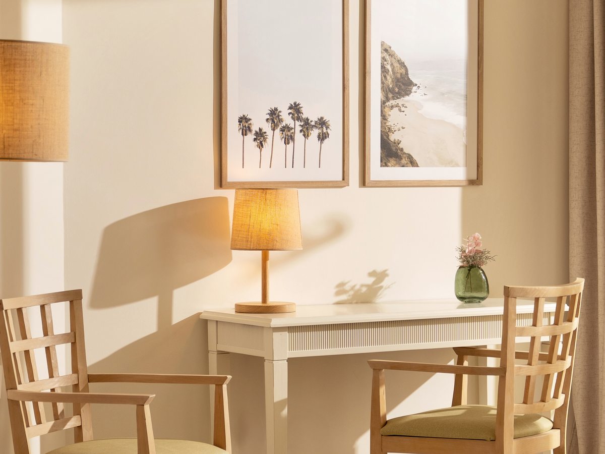 A corner of a guest room with two wooden chairs, a small writing desk, a table lamp and framed coastal prints on a neutral wall.