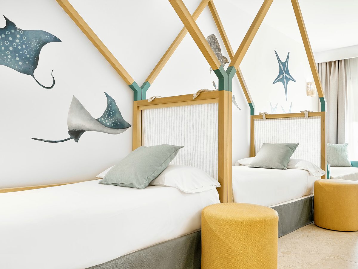 A children's bedroom with two single beds under house-frame timber headboards, manta-ray wall art and a yellow stool by the window.