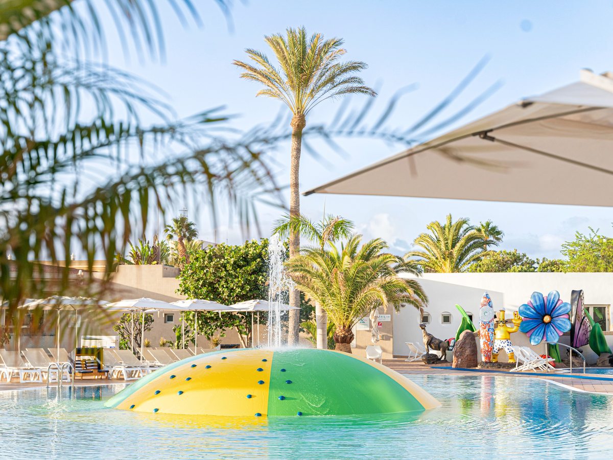 A shallow children's pool with a green and yellow inflatable dome spraying water, palm trees and white parasols around the pool terrace.