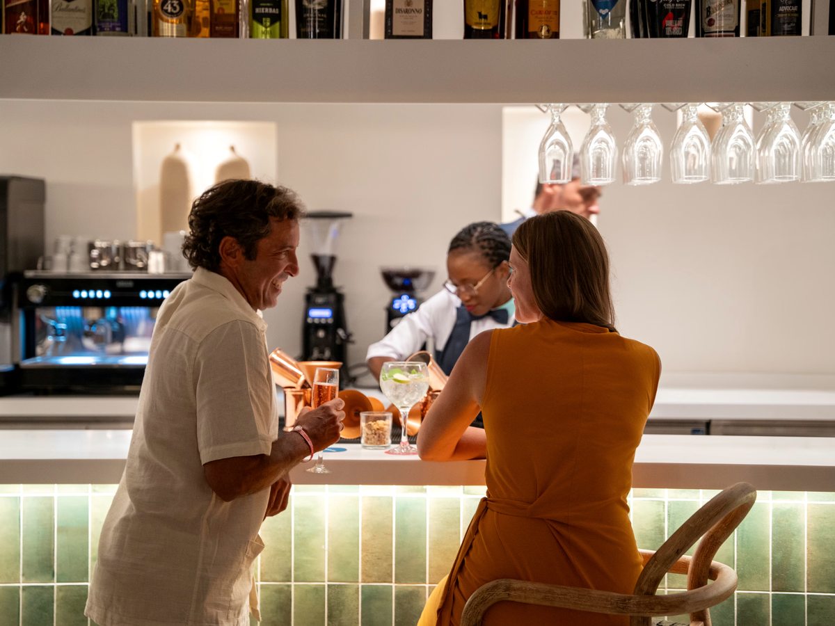A bartender behind a green-tiled bar counter serving two seated guests, with coffee machines and wine glasses along the back shelf.