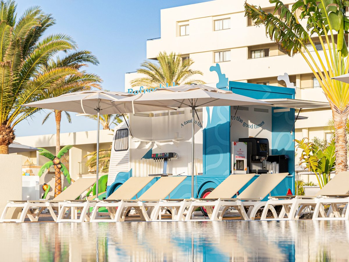 A blue food-truck-style snack bar beside the pool, with sun loungers, white parasols and palm trees around a pool terrace.