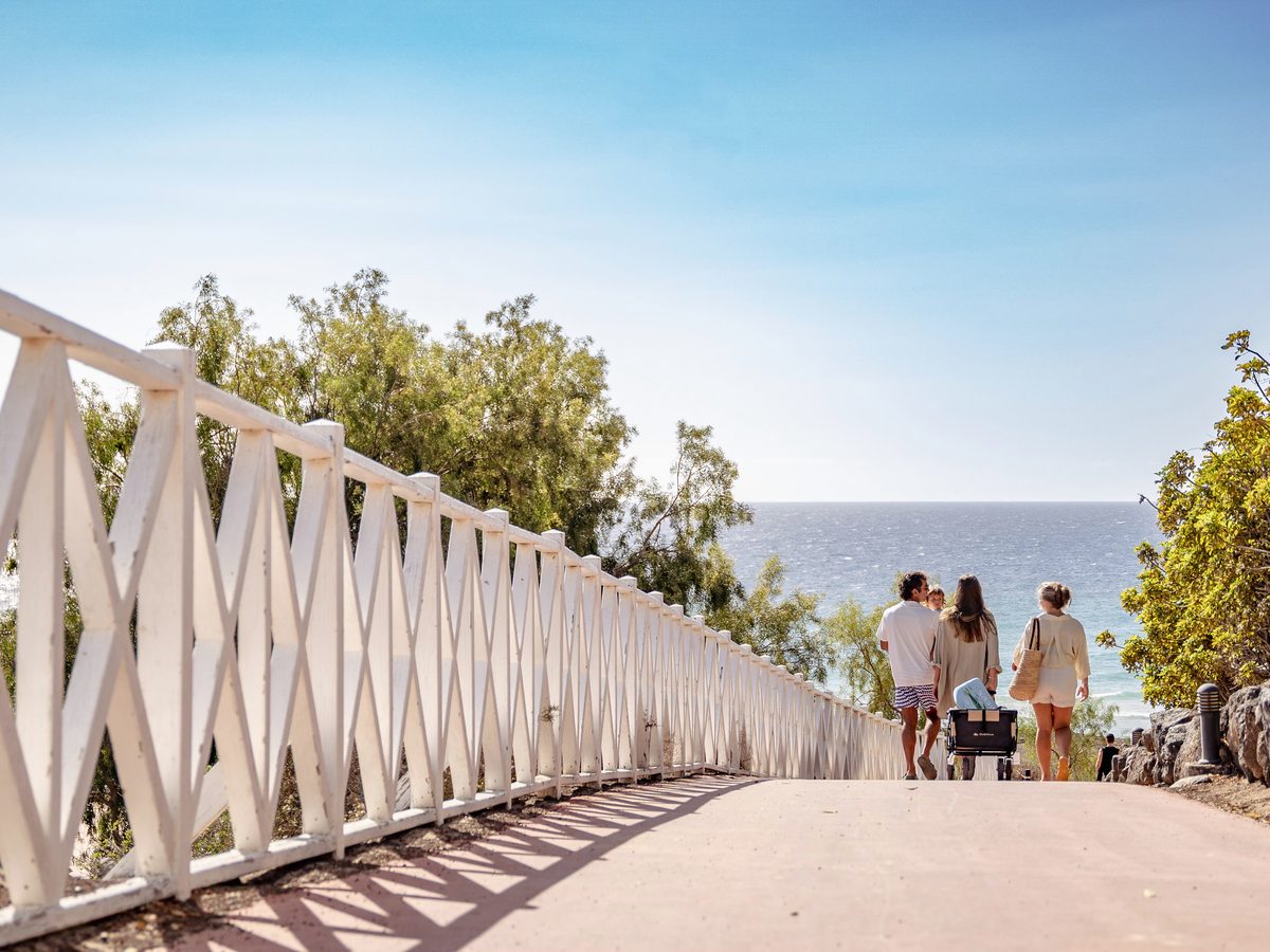 A wide paved path with white railings sloping downhill towards the sea, a family walking down it pushing a pushchair under a clear blue sky.