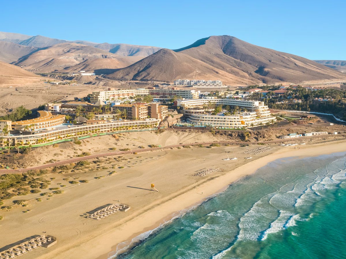 An aerial view of the wide sandy beach and breaking waves, with the seafront resort buildings on the left and bare volcanic hills rising behind.