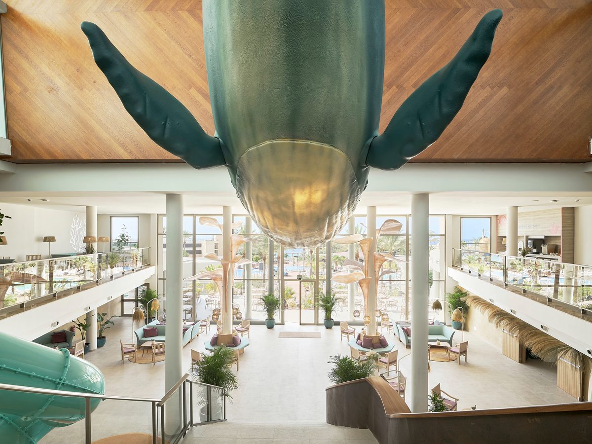 A bright double-height lobby atrium with a large green whale sculpture suspended from the wood-panelled ceiling, balcony walkways, indoor plants and lounge seating.