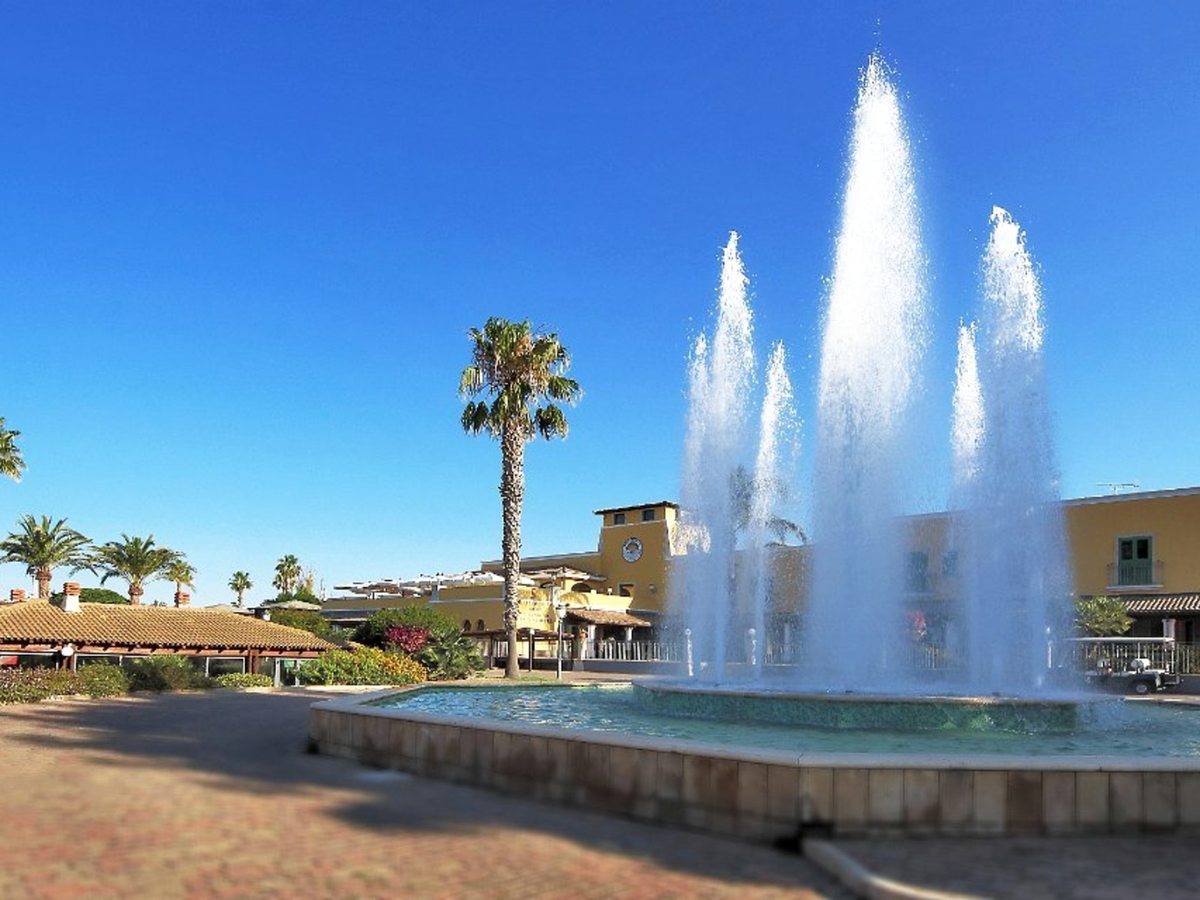 An ornamental fountain sending up tall water jets from a circular basin on a paved plaza, ringed by palms and low resort buildings.