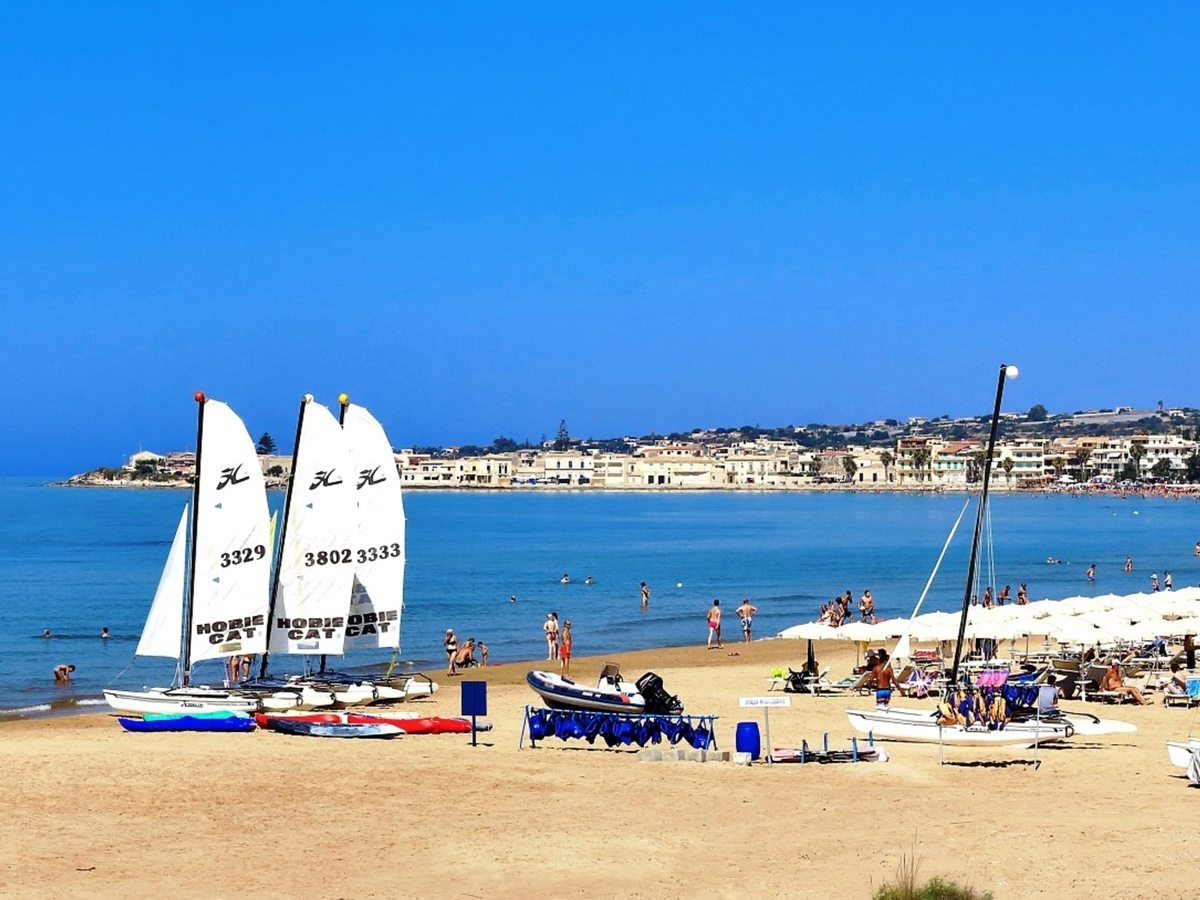 Small sailing dinghies drawn up on the sand with beachgoers, a bay and a coastal town across the water.