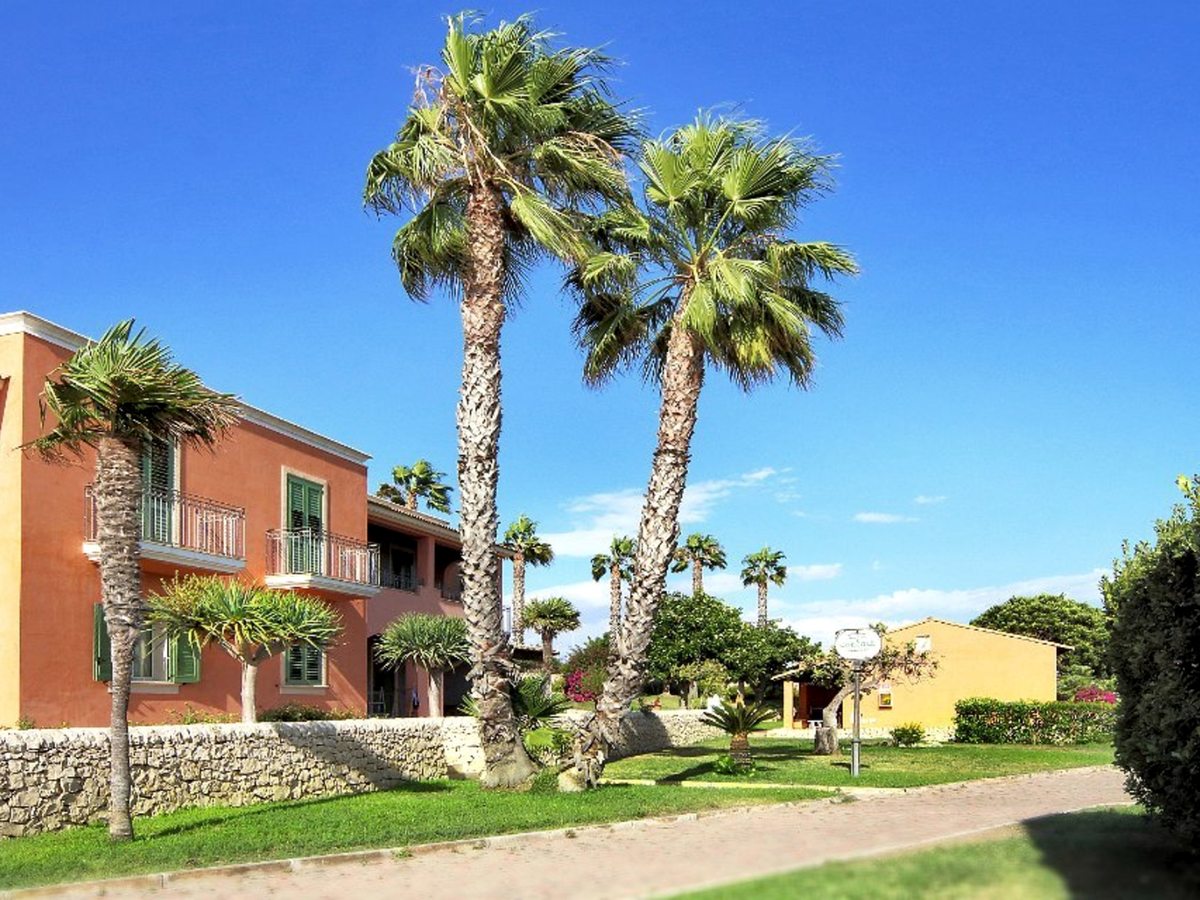 Two-storey pastel accommodation blocks with upper-floor balconies among tall palms and a lawn behind a low stone wall.