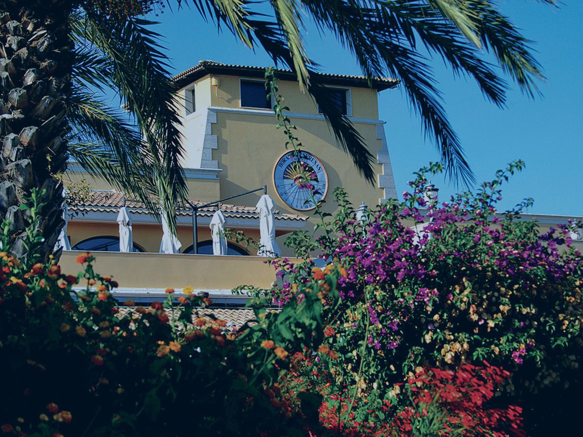 A pale-yellow building framed by palms and beds of pink bougainvillea in full flower under a clear sky.