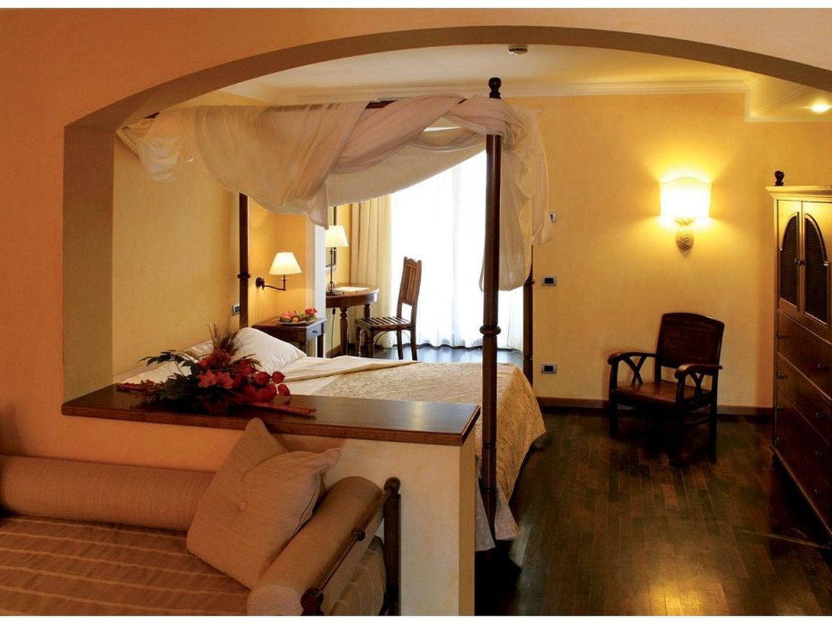 A suite with a canopied bed in an arched alcove and a separate sitting area with a sofa and armchair under warm lighting.