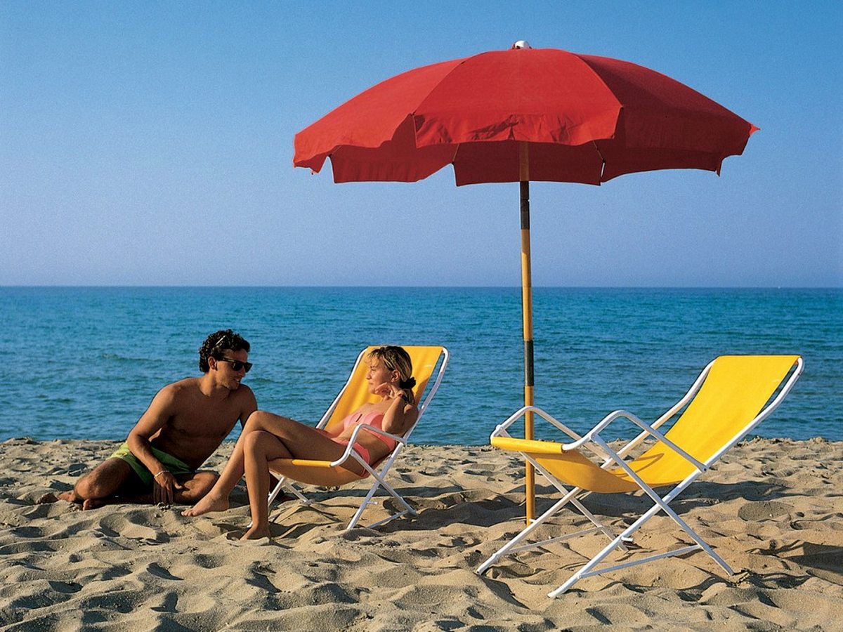 A fine-sand beach with a red parasol and yellow deckchairs, a couple relaxing and calm blue sea beyond.