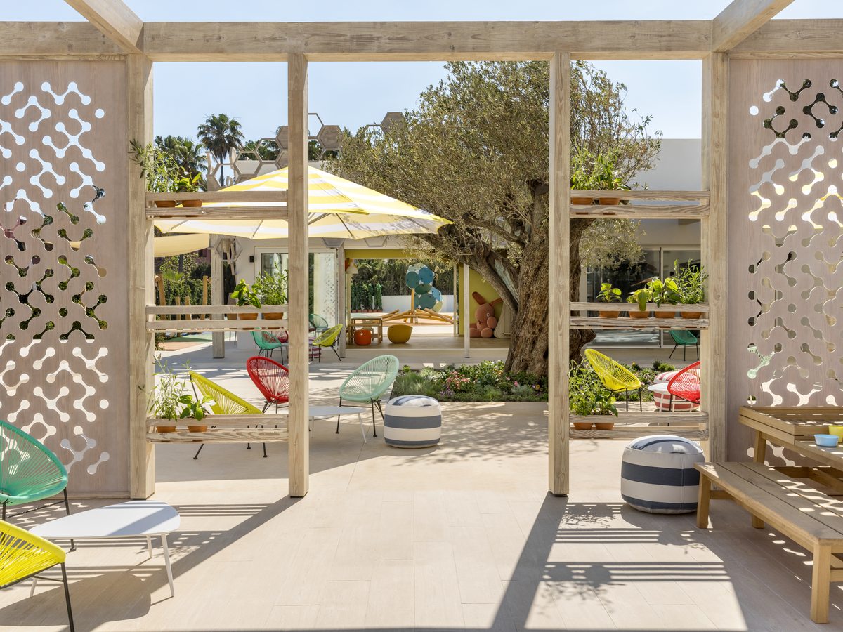 The Heroes Village at Ikos Andalusia, a shaded outdoor terrace under a timber pergola with children’s chairs, low tables and benches among the trees.