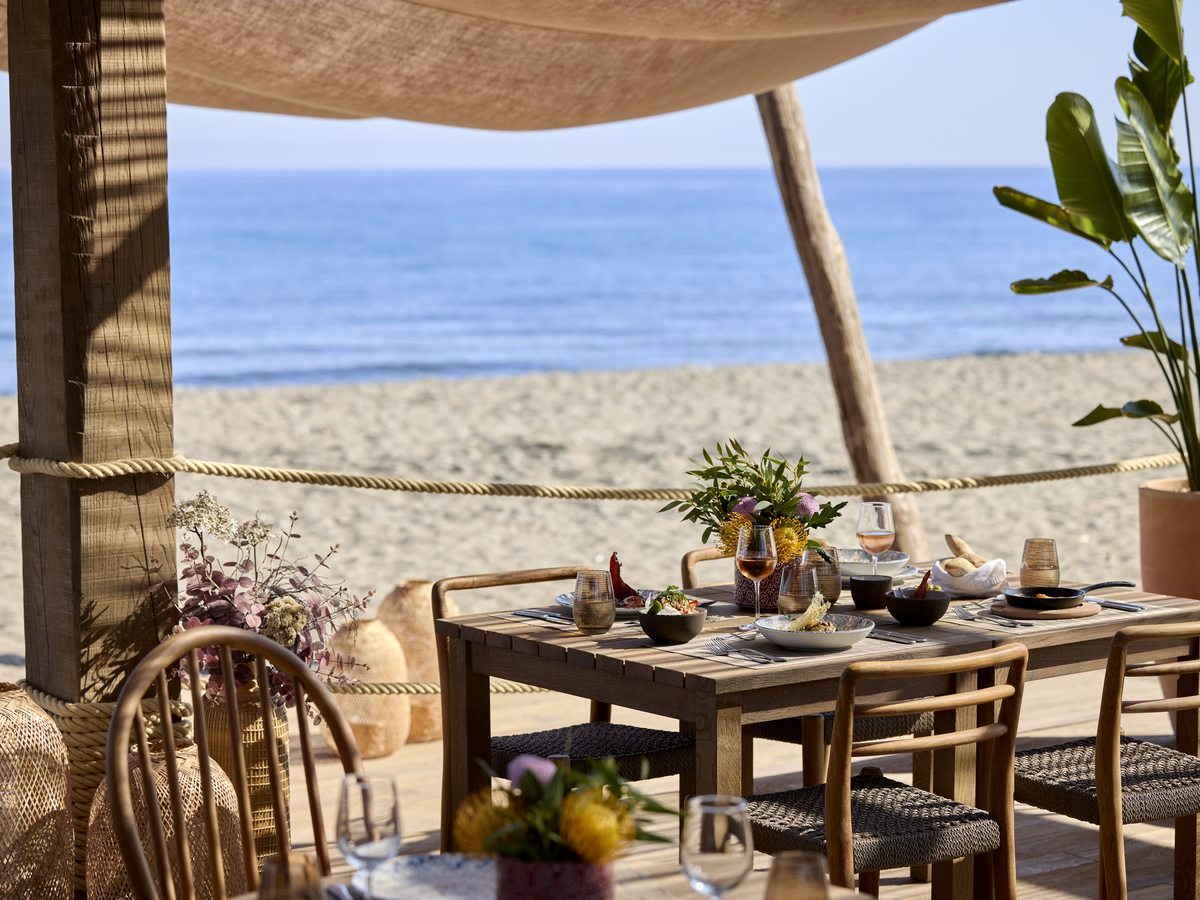 A laid table under the canopy at Chiringuito, the beachfront restaurant at Ikos Andalusia, with the sand and the sea in front of it.