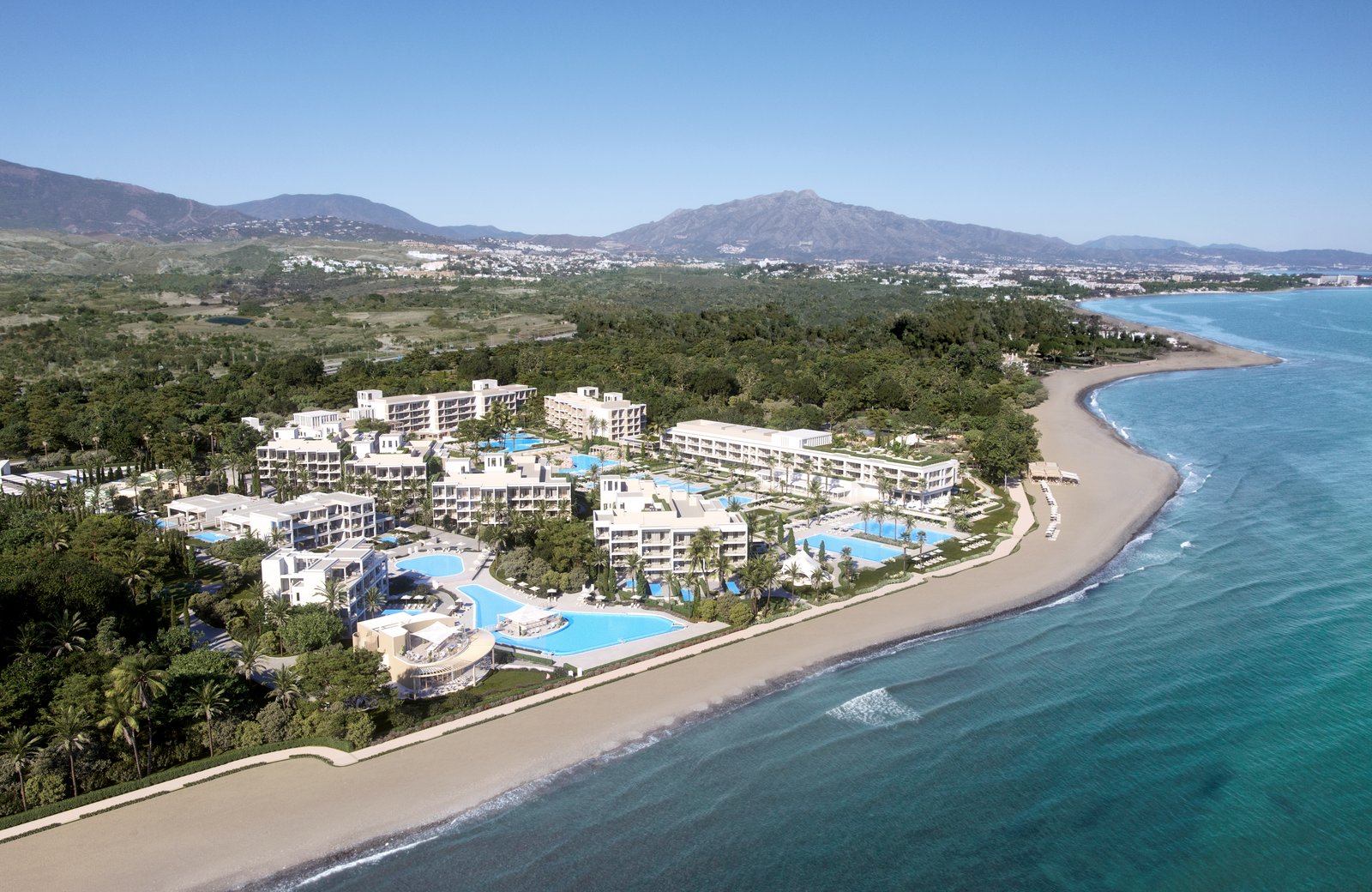 Ikos Andalusia from the air: a low white seafront resort set in palm gardens along a curving bay, with hills behind.