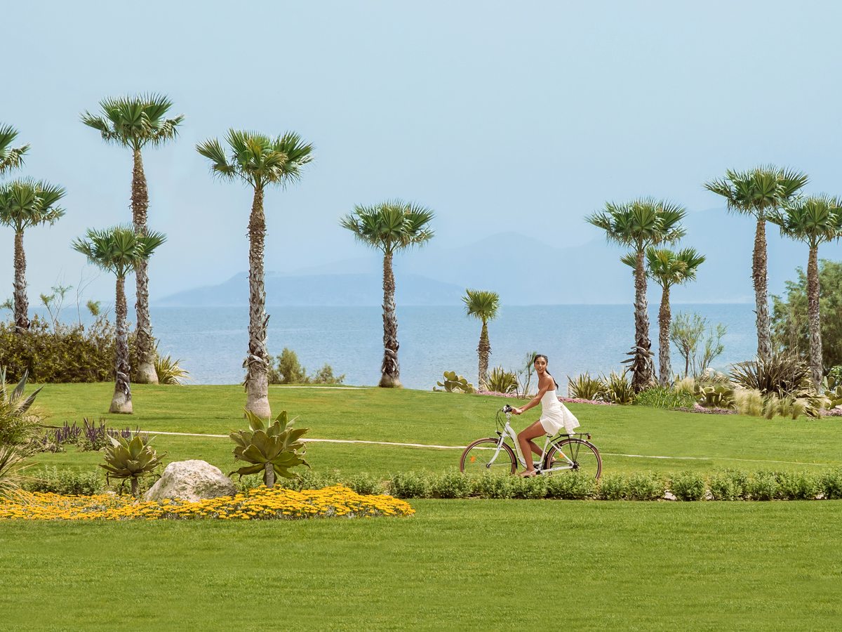 A guest cycling a path across the resort lawns, with palms, flowering borders and the hills beyond.