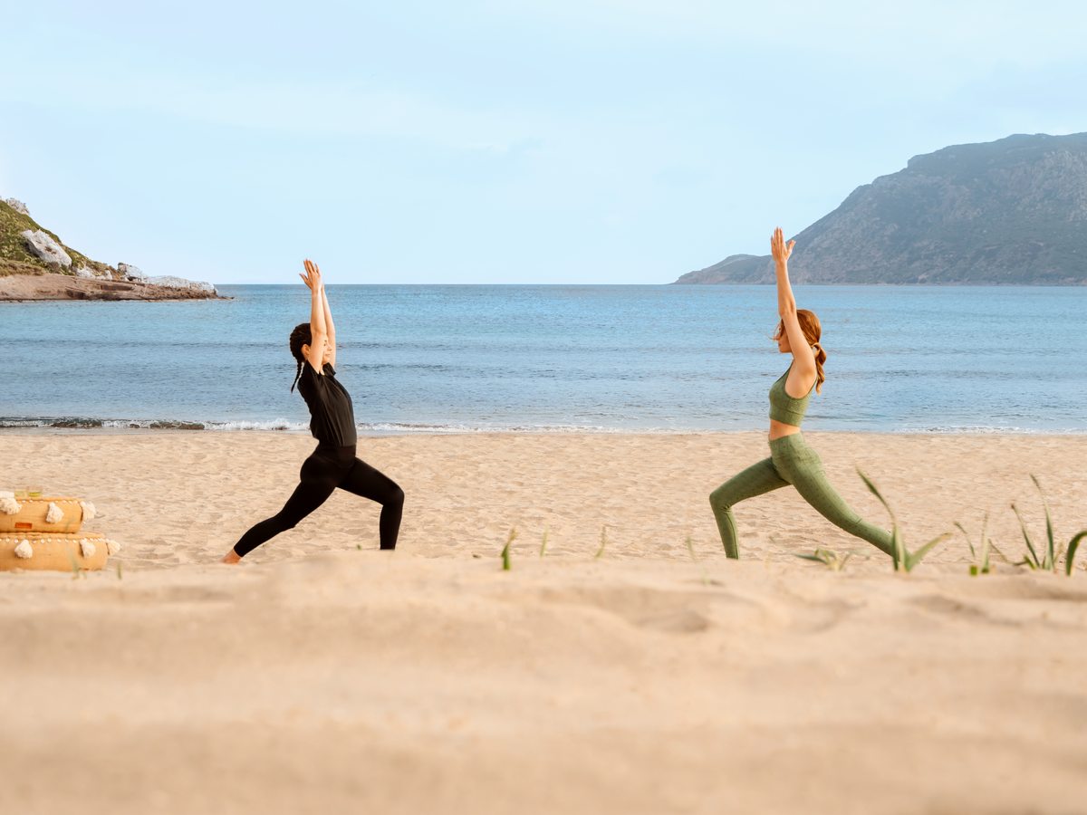 Two people holding a yoga pose on the sand at the water's edge, with the headland across the bay behind them.
