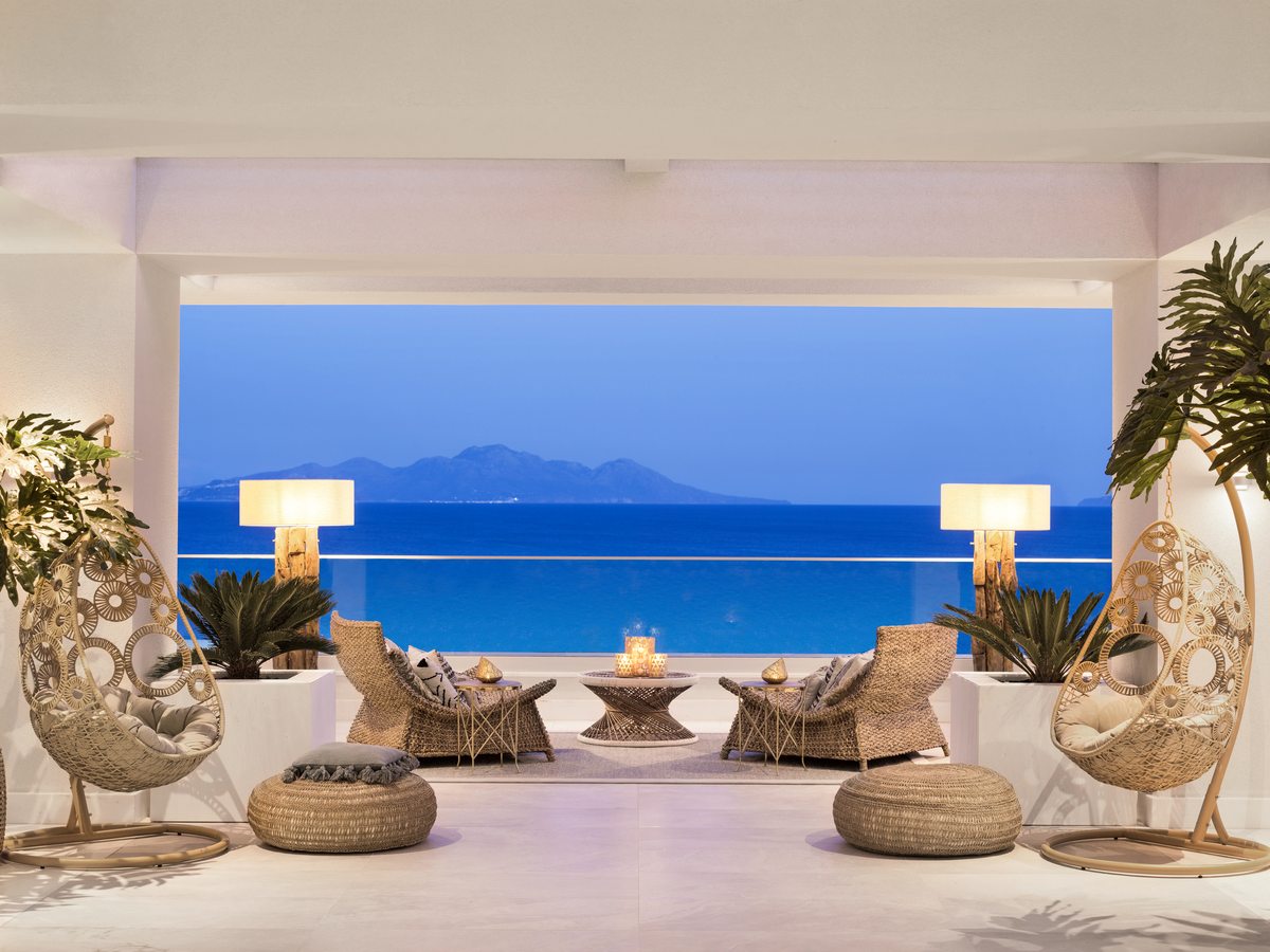 An open-sided veranda with hanging rattan chairs, low seating and floor cushions, framing the sea and the hills beyond.