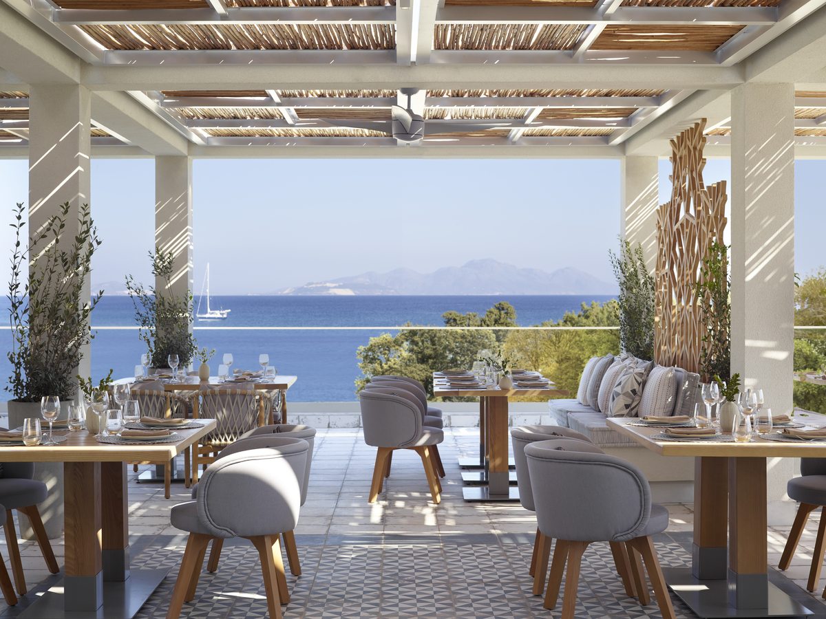 The dining room at Kos, open along one side to a terrace and the sea, with dark wooden chairs and stone-clad columns.