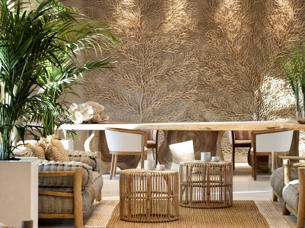 The lobby at Ikos Aria, a pale open room with cane armchairs, a long wooden table and a large woven wall panel behind it.