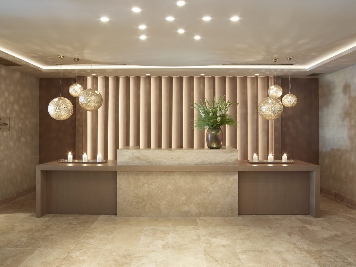 Spa reception desk in warm stone, with slatted timber panelling and hanging globe lights.
