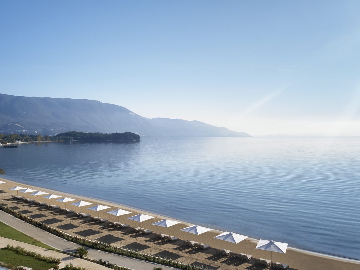 Rows of parasols and sunbeds along the shore, the calm sea and a green headland beyond.