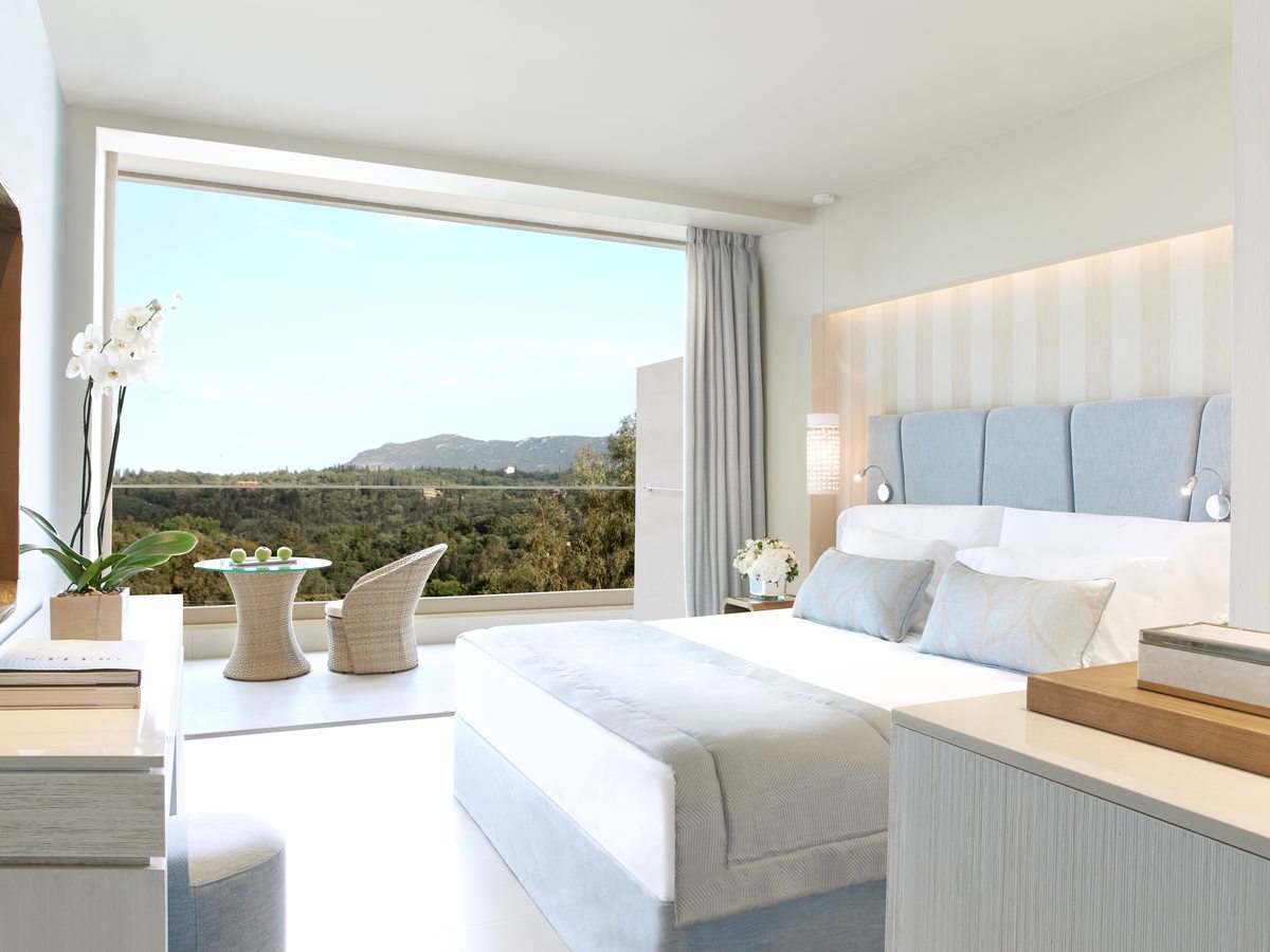 A double bed with a padded blue headboard beside glass doors open to a terrace and the wooded hills.