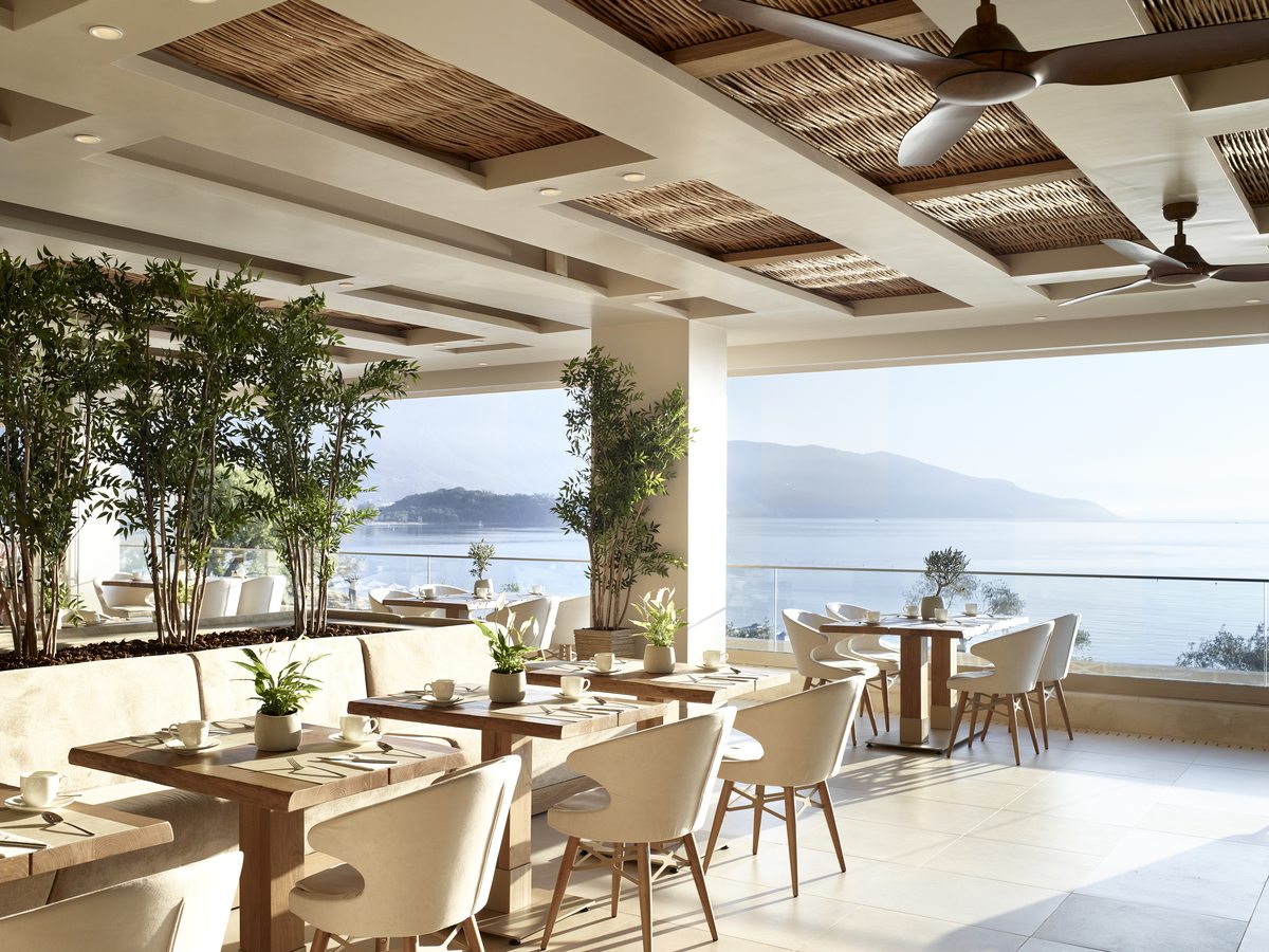 Dining room with pale tables and rattan chairs under a slatted ceiling, open along one side to the sea.