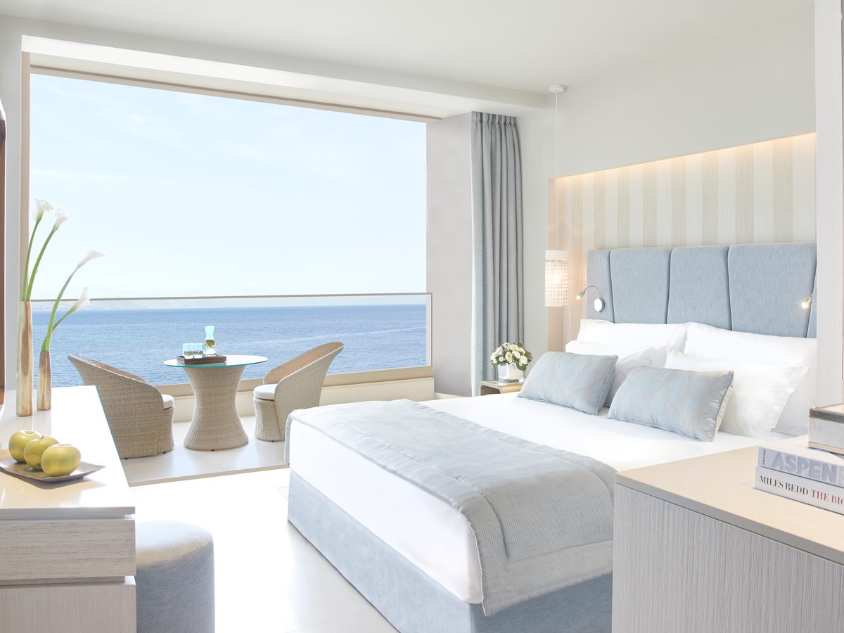 Bedroom with a wide bed and a padded headboard, glass doors open to a terrace table and the sea.