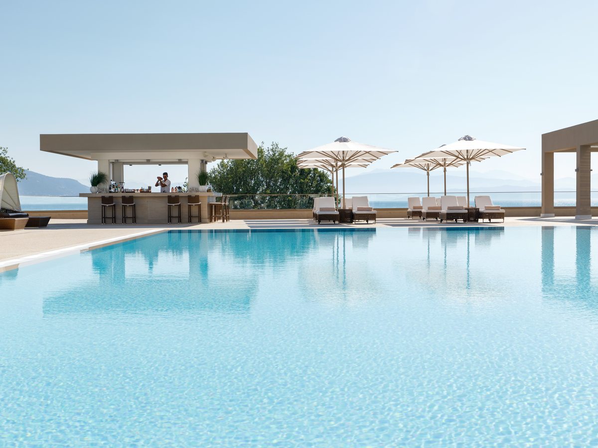 Poolside bar under a flat canopy, with parasols and sun loungers around the pool edge and the sea behind.