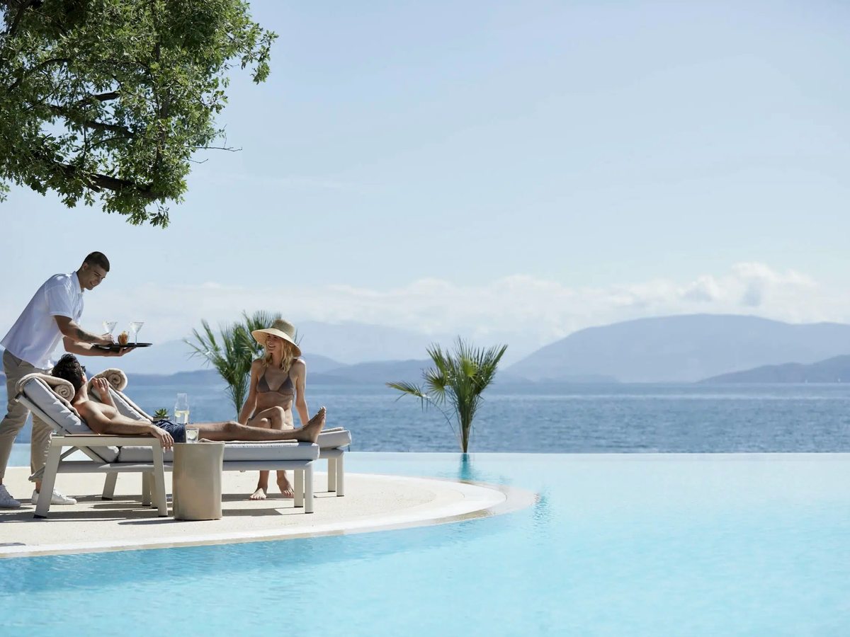 An infinity pool with sun loungers on a shaded terrace at its edge, the calm sea and distant mountains beyond.
