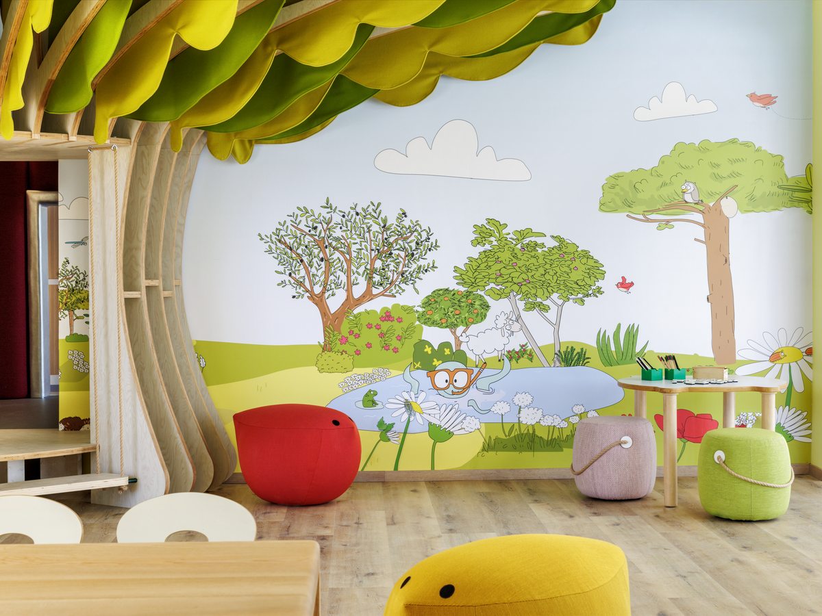 A kids club playroom at an Ikos resort, with a painted woodland mural, soft seats and low tables.