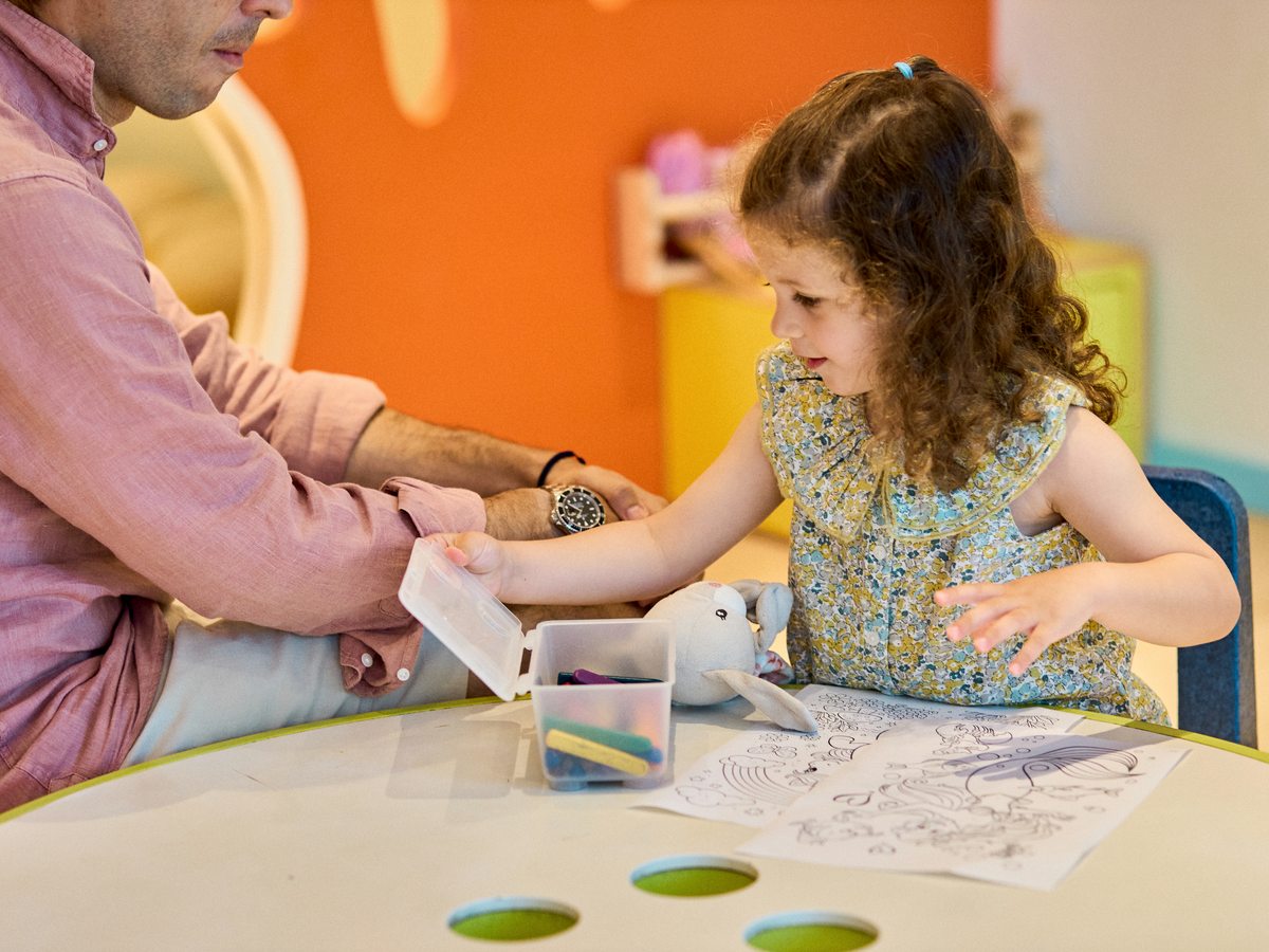 A young child colouring a picture at a kids club table at an Ikos resort, with a grown-up sitting beside her.