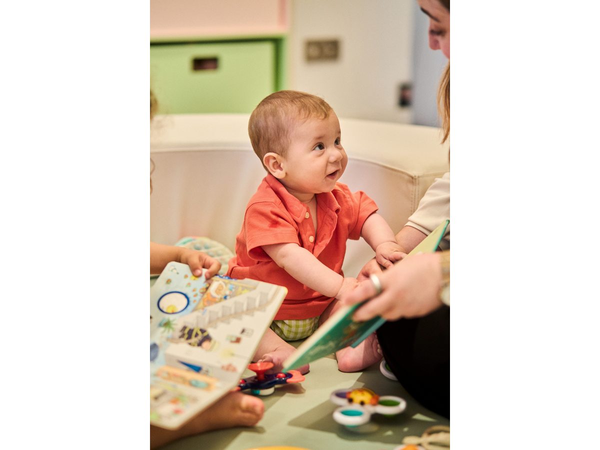 A baby sitting on the floor of a playroom at an Ikos resort with wooden toys and a book, a carer beside him.
