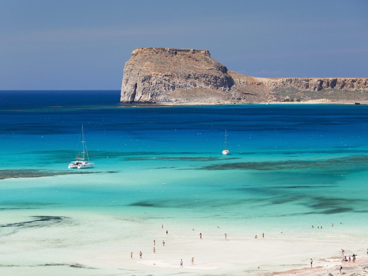 Balos lagoon in western Crete, shallow turquoise water over pale sand below the Gramvousa headland, with two yachts anchored offshore.