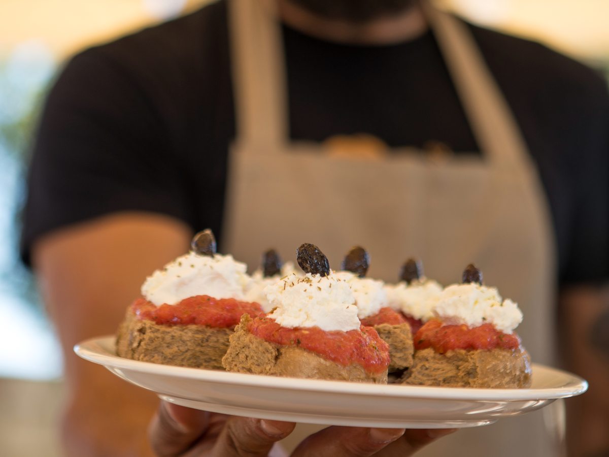 A chef in an apron holding out a plate of barley rusks topped with tomato, soft white cheese and olives.