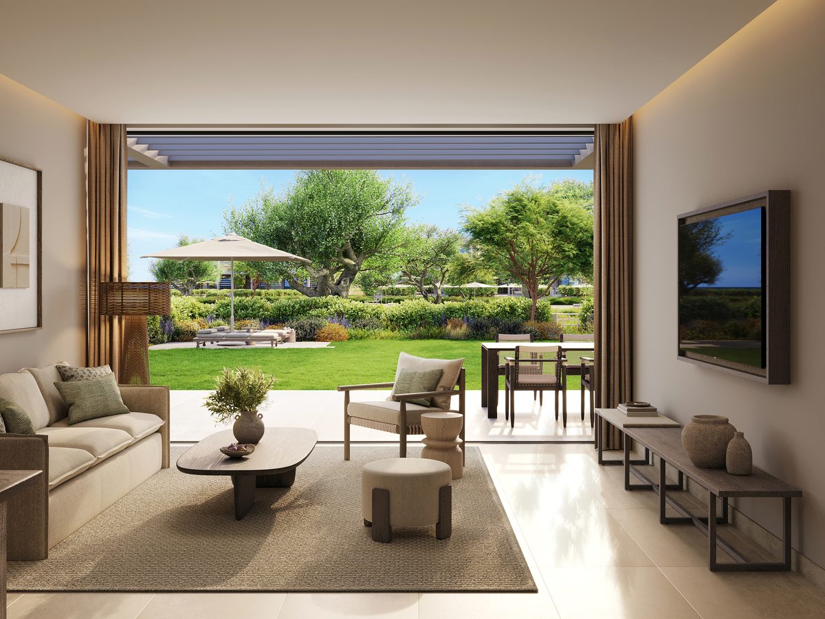 The lounge of a Deluxe Two Bedroom Suite at Ikos Kissamos, with a sofa, a dining table and a wall of glass folded back onto a lawn and garden.