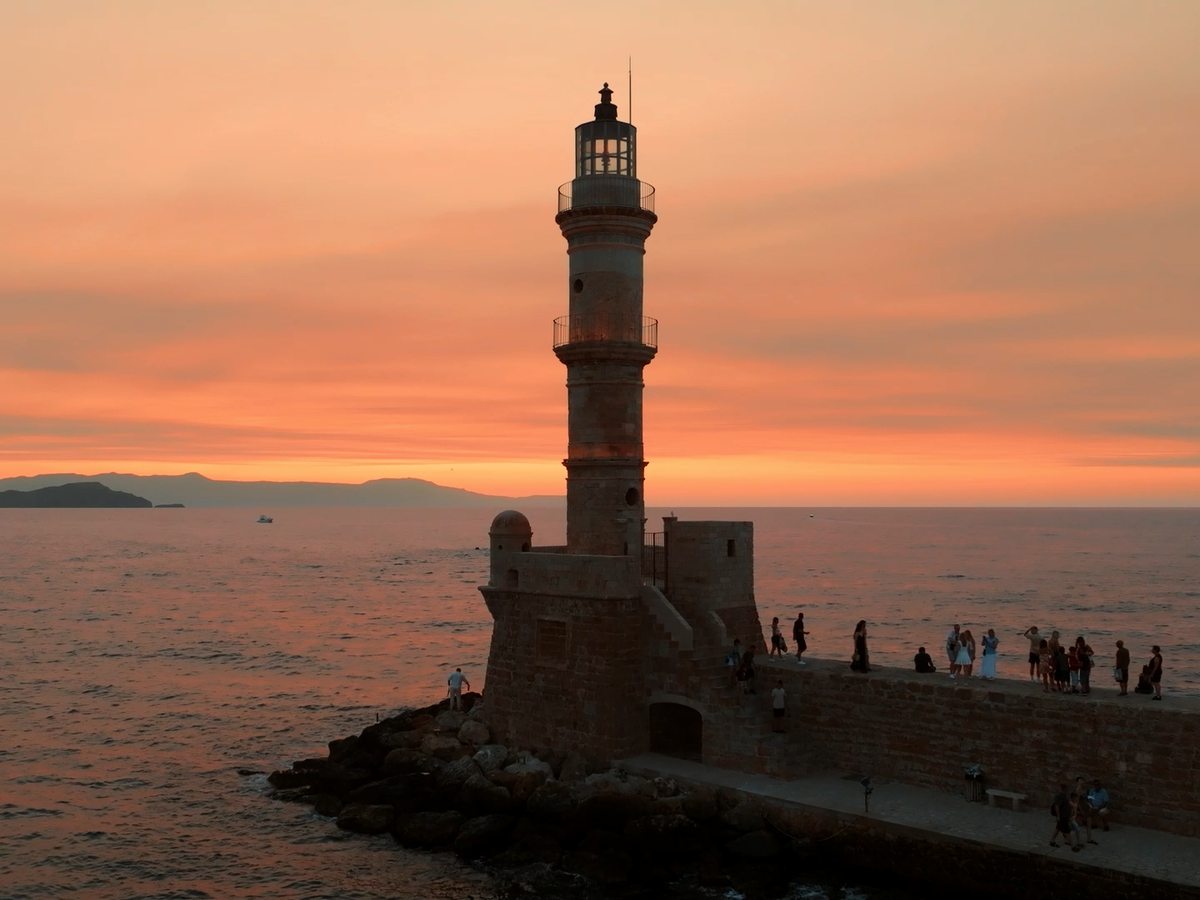 The Venetian lighthouse at the entrance to Chania harbour at sunset, with people along the harbour wall.