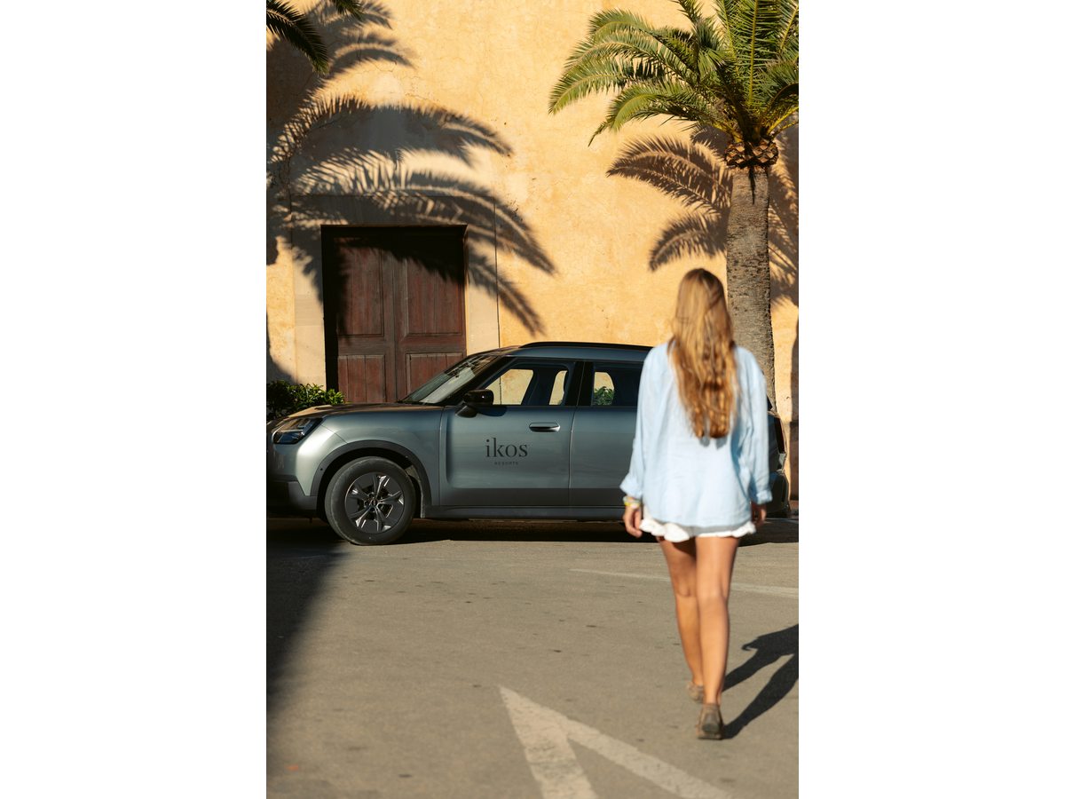 A small car with an Ikos badge on the door, parked beside a stone building and palm trees, with a guest walking towards it.