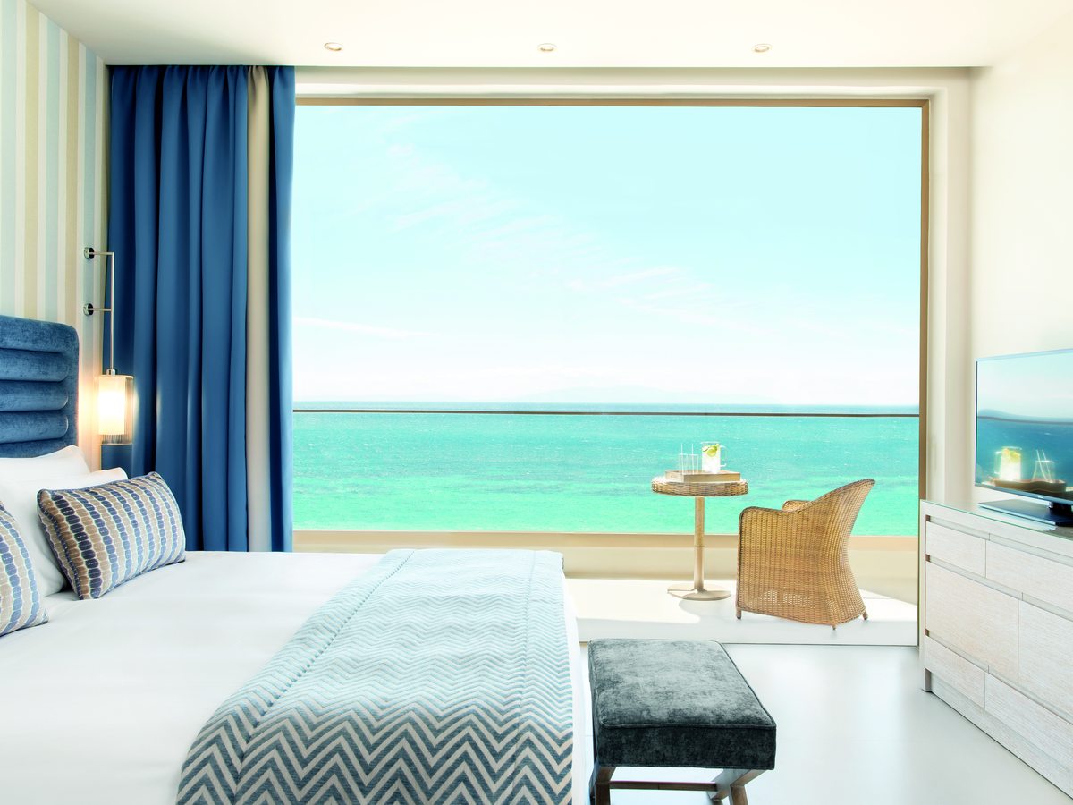 A Superior Double Room with a king bed facing a full-width window onto turquoise sea, and a wicker chair and table on the balcony.