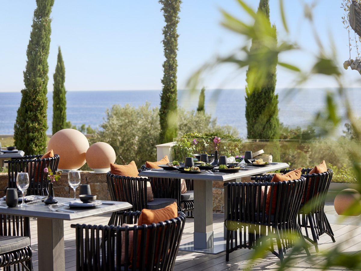 The outdoor deck at Anaya, with dark tables, orange cushions and round lanterns, cypresses and the sea behind.