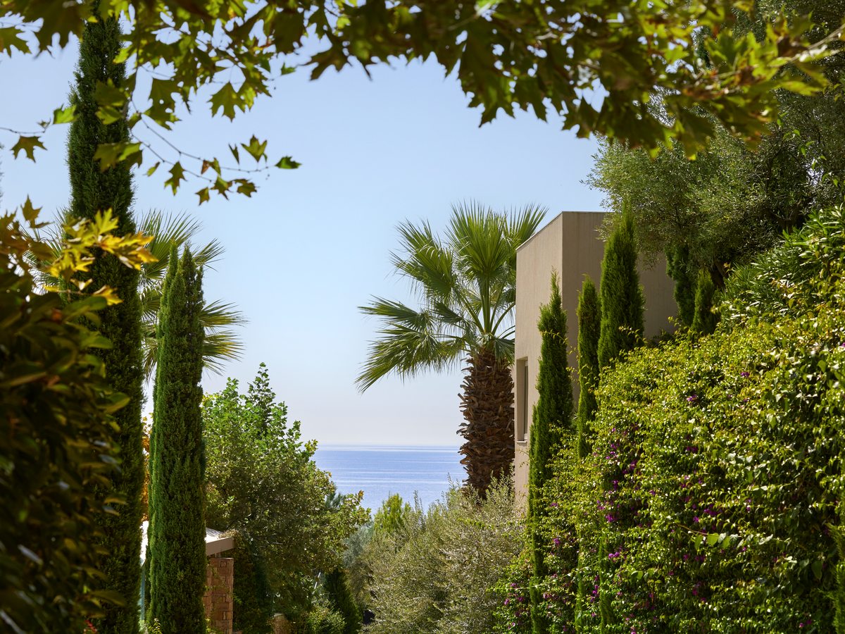 A white resort building set among cypresses, palms and plane trees, with a band of blue sea beyond the greenery.
