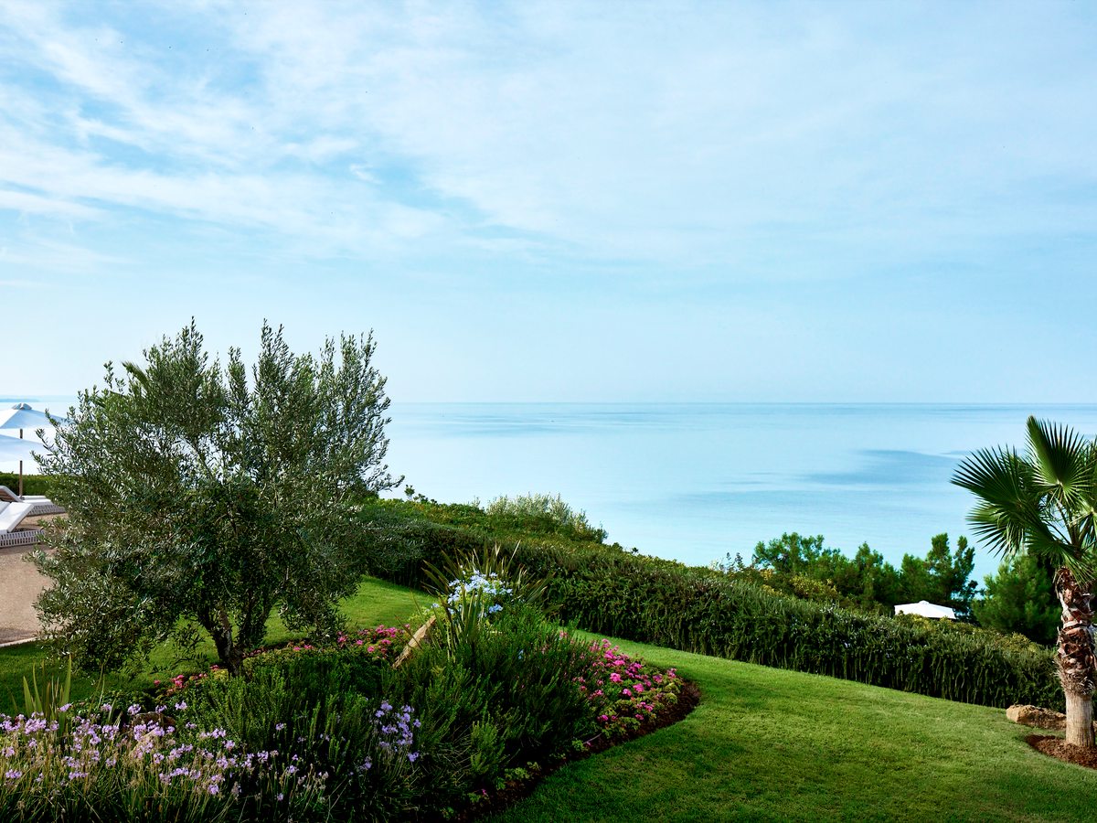 Terraced lawns and planted borders with an olive tree and a palm, running down past clipped hedges to the calm blue Aegean.
